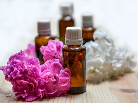 The image shows small brown bottles with white caps, surrounded by pink and white flowers, suggesting an arrangement of essential oils and blossoms.