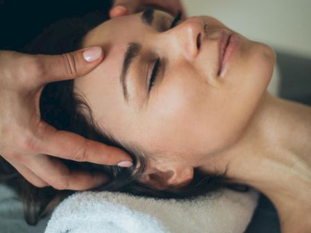 A person is lying down with their head on a towel, receiving a relaxing facial massage with closed eyes and a content expression.