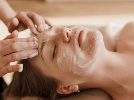 A person is receiving a facial treatment with a cream mask being applied to their face by another person, likely in a spa or beauty setting.