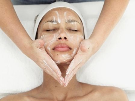 A person receiving a facial treatment, with hands applying a cream or mask on their face, while lying down on a white towel.