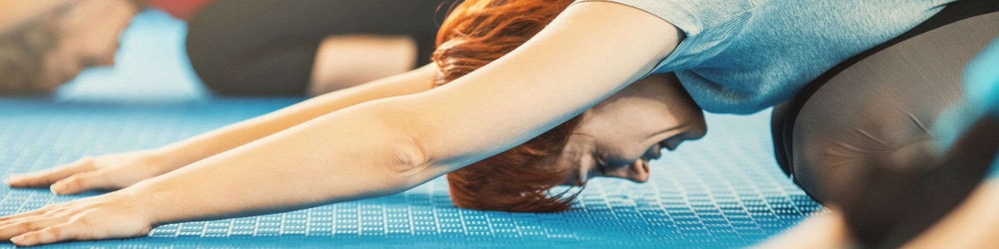 People in a yoga class practicing a child's pose on blue mats indoors, focused and stretching forward with their arms extended.