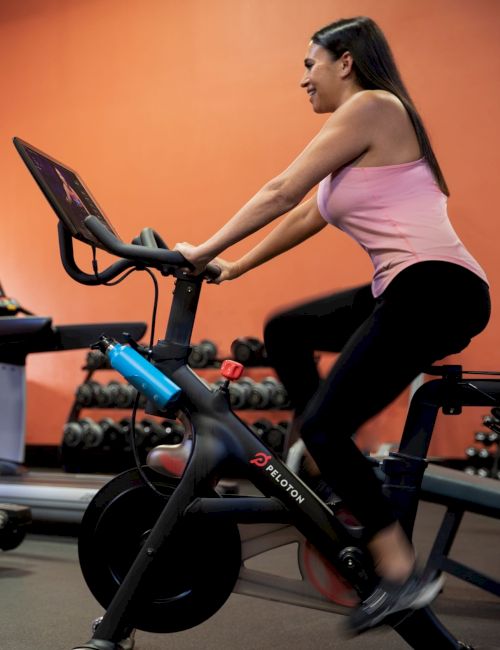 A person in a pink top riding a stationary bike at the gym, with a screen in front of them and dumbbells in the background.