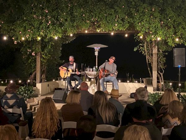 Two musicians are performing on a small outdoor stage surrounded by greenery, with an audience seated and watching.