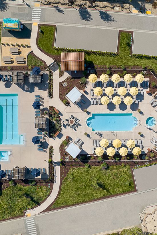 This image depicts an aerial view of a pool facility with two pools, multiple umbrellas, lounging areas, pathways, and surrounding greenery.