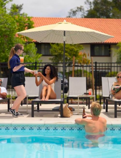 Several people relax by a pool, with some sunbathing on lounge chairs and one person in the pool. A server is handing a drink to a woman.