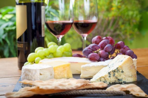 The image shows a wine and cheese platter with different cheeses, green and red grapes, and crackers, accompanied by two glasses of red wine.