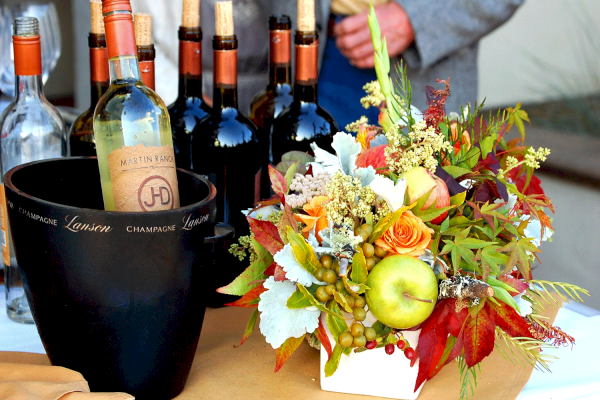 The image shows multiple wine bottles, a black ice bucket, and a colorful floral arrangement with fruits and flowers on a table.