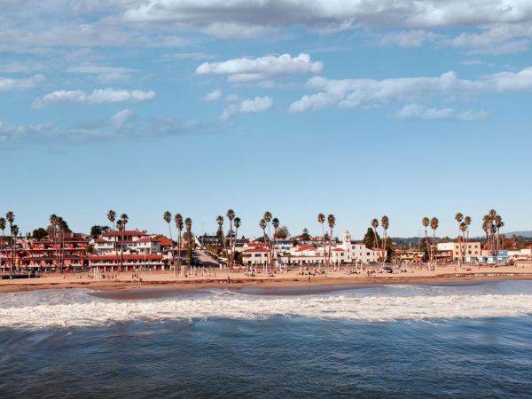 A coastal scene with waves hitting the sandy shore, lined with palm trees and buildings in the background under a cloudy sky.