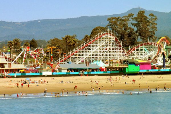 The image shows a beachfront amusement park with roller coasters, colorful buildings, and people enjoying the beach and water.