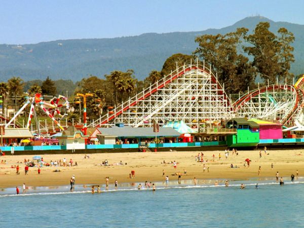 The image shows a beachfront amusement park with roller coasters, colorful buildings, and people enjoying the beach and water.