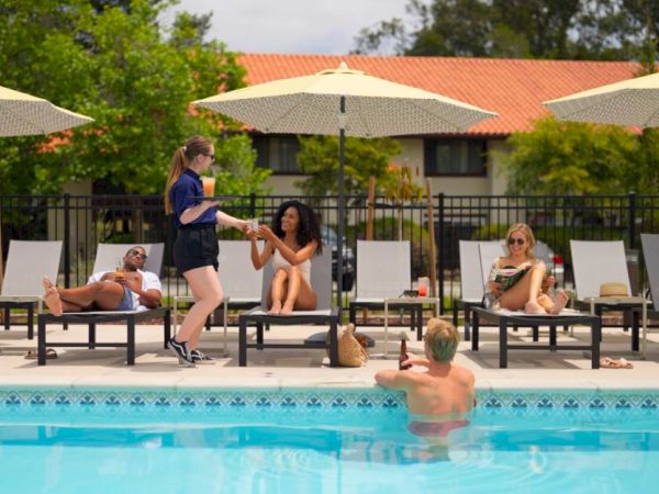People relaxing by a pool with a few on lounge chairs under umbrellas, and one person standing, serving drinks to the others.