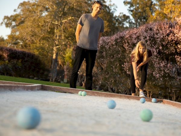 Two people are playing bocce ball on an outdoor court surrounded by trees and bushes in a sunny setting.