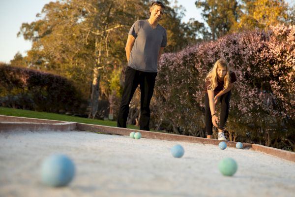 Two people outdoor playing bocce ball on a dirt court. The background has greenery.