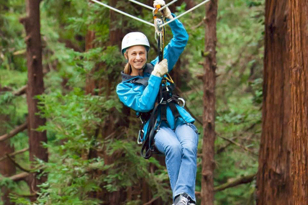 A person wearing a helmet and harness is zip-lining through a forested area with tall trees and greenery all around.