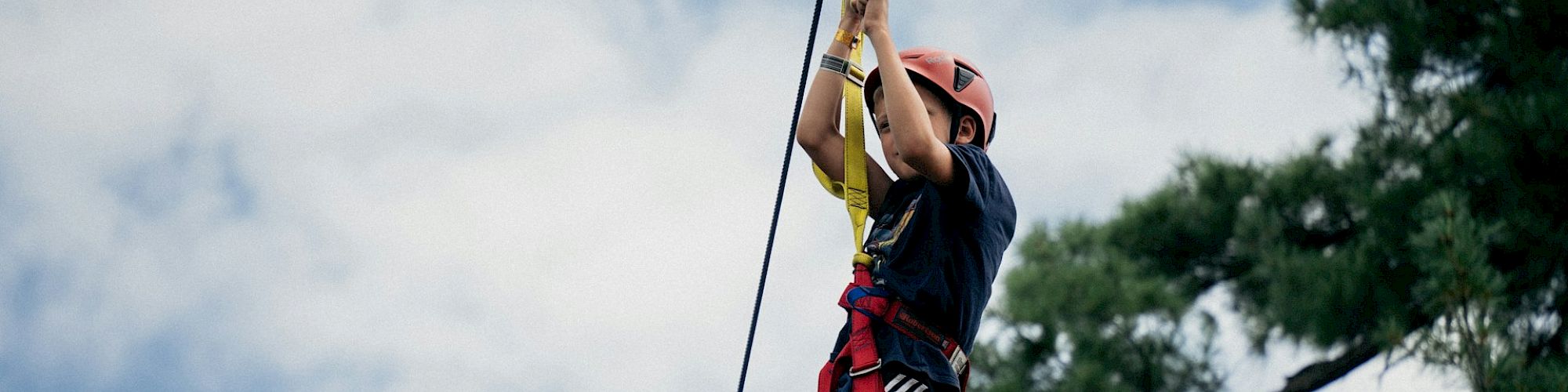 A person wearing a helmet and safety gear is zip-lining through the trees, with a cloudy sky in the background.