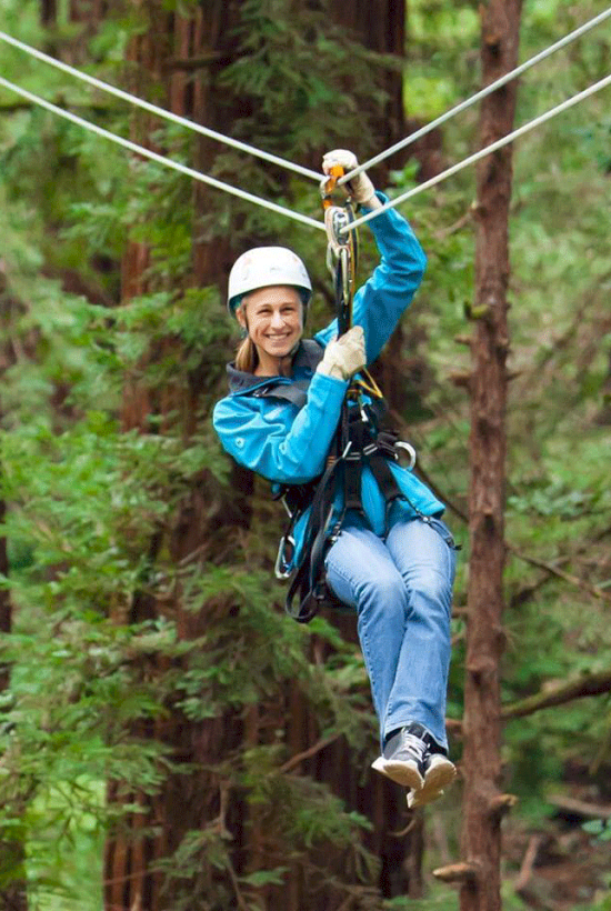 A person is zip-lining through a forested area, wearing a helmet and safety gear, looking happy and enjoying the adventure.