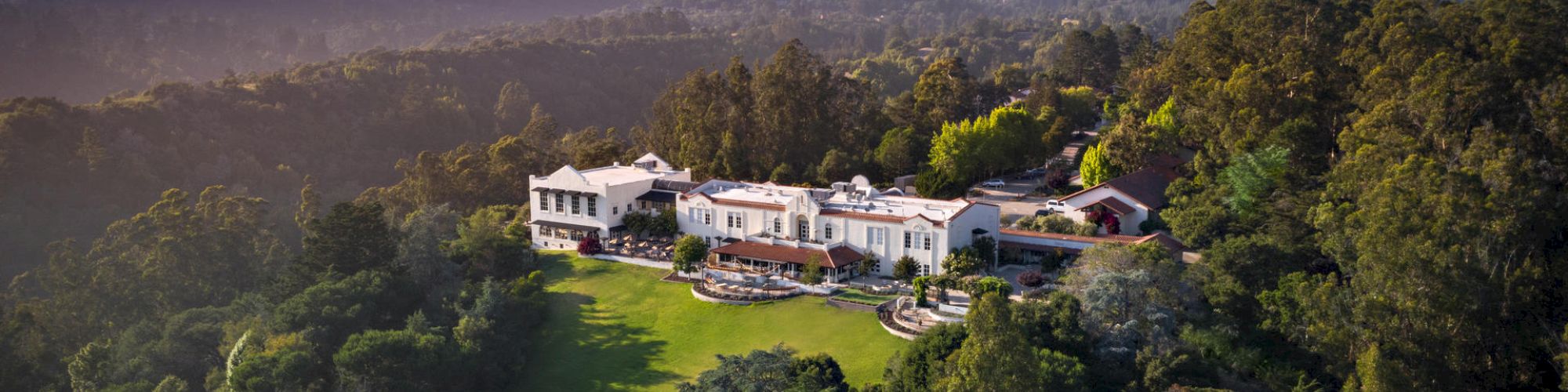 An aerial view of a large mansion surrounded by dense green forest and hills under a clear sky.