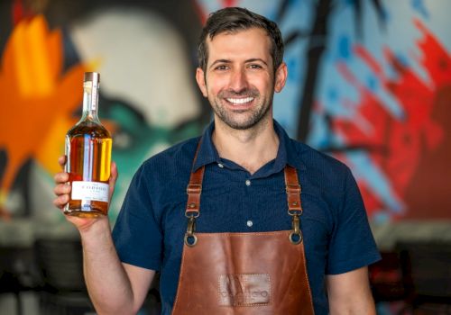 A man wearing a leather apron holds a bottle of liquor, standing in front of a colorful graffiti backdrop, smiling at the camera.