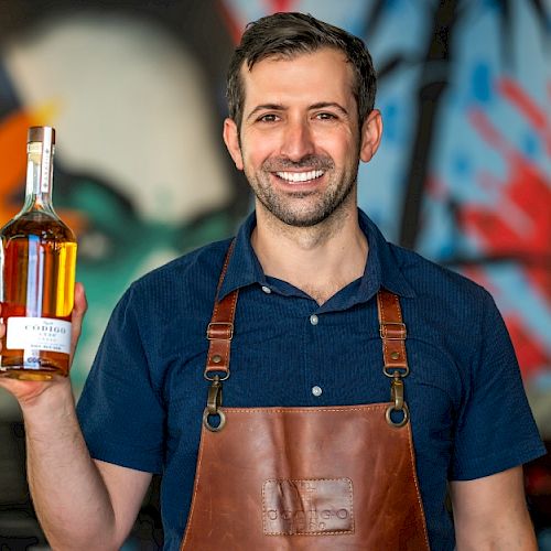 A man wearing a leather apron holds a bottle of liquor, standing in front of a colorful graffiti backdrop, smiling at the camera.