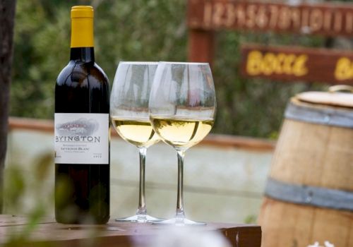 A bottle of white wine is beside two filled wine glasses on a wooden surface, with a barrel and signs in the background.