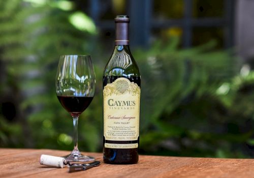 The image shows a bottle of Caymus Vineyards Cabernet Sauvignon, a filled wine glass, a corkscrew, and a cork on a wooden table.