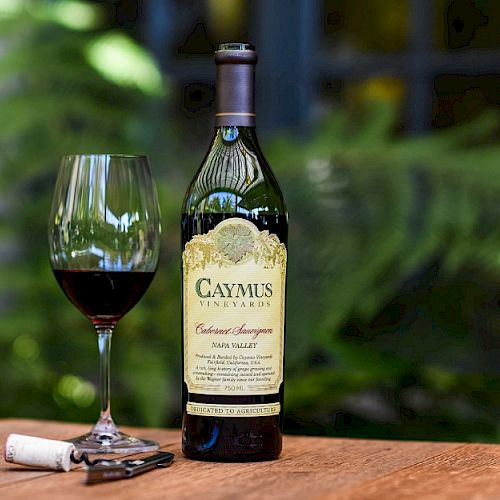 The image shows a bottle of Caymus Vineyards Cabernet Sauvignon, a filled wine glass, a corkscrew, and a cork on a wooden table.