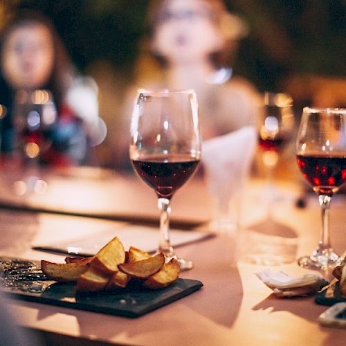 The image shows a table with several glasses of red wine, plates of food, and people in the background, slightly out of focus.