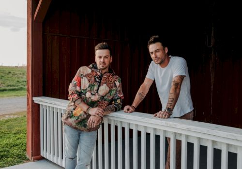 Two men standing on a porch, leaning against a white railing with wooden boards in the background and green grass in the distance, ending the sentence.