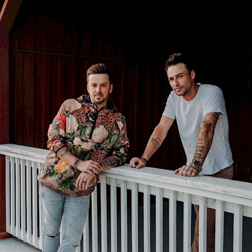 Two men standing on a porch, leaning against a white railing with wooden boards in the background and green grass in the distance, ending the sentence.