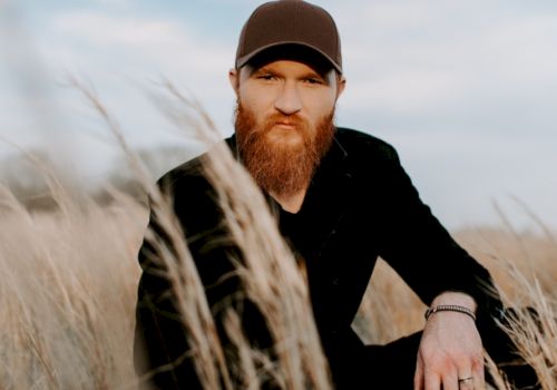 A man with a beard and wearing a cap is sitting in a grassy field under a partly cloudy sky, looking directly at the camera.