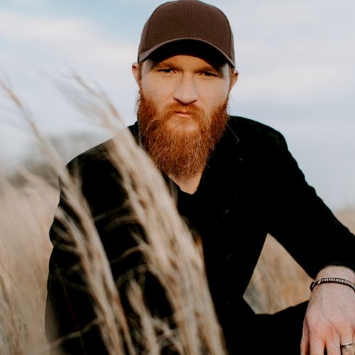 A man with a beard and wearing a cap is sitting in a grassy field under a partly cloudy sky, looking directly at the camera.