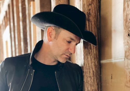 A person wearing a black cowboy hat and jacket stands thoughtfully against wooden posts in a rustic setting.