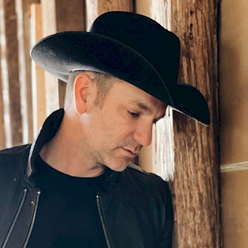 A person wearing a black cowboy hat and jacket stands thoughtfully against wooden posts in a rustic setting.