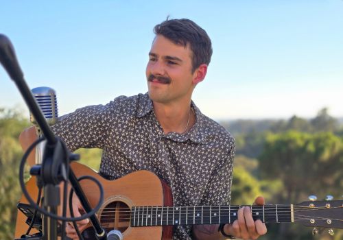 A person playing an acoustic guitar outdoors near a microphone, with a scenic background of trees under a clear sky ending the sentence.