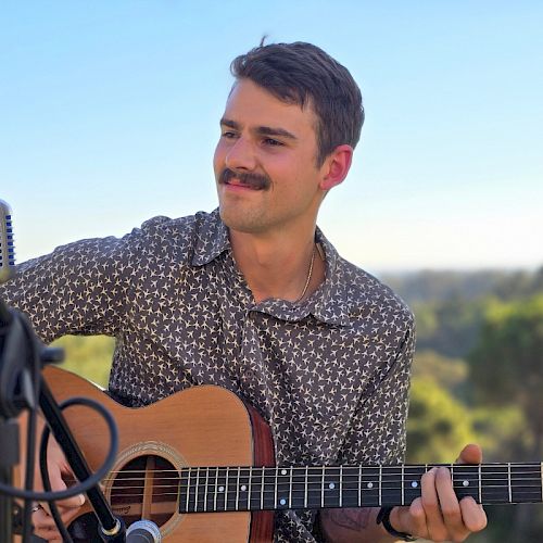 A person playing an acoustic guitar outdoors near a microphone, with a scenic background of trees under a clear sky ending the sentence.