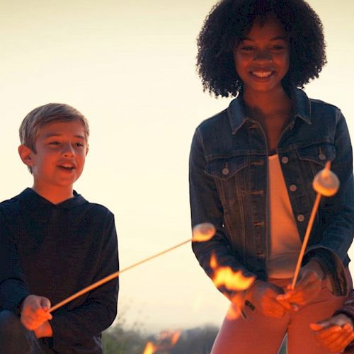 A group of four people are gathered around a campfire, smiling and roasting marshmallows on sticks.