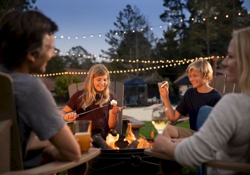 A family enjoys roasting marshmallows and making s'mores around a campfire in their backyard, with string lights in the background.