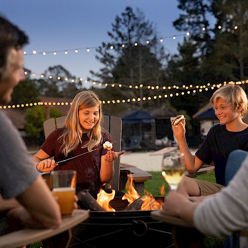 A family enjoys roasting marshmallows and making s'mores around a campfire in their backyard, with string lights in the background.