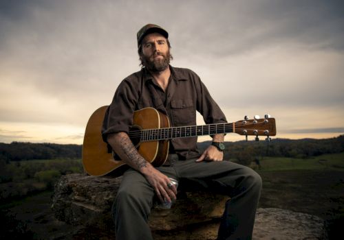 A man sits on a rock holding an acoustic guitar and a beverage can, with a vast outdoor landscape in the background, under a cloudy sky.