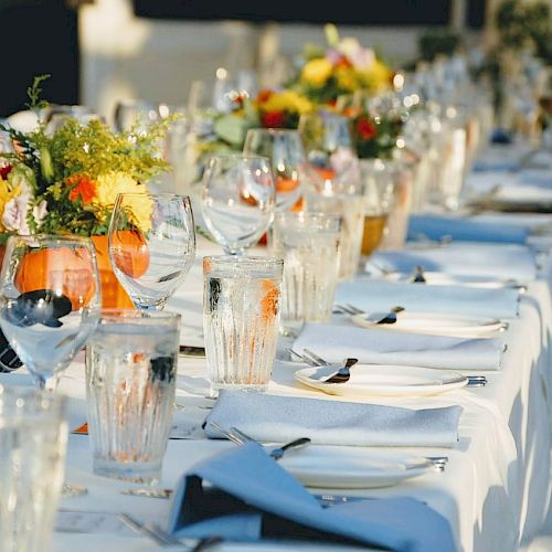 A long, elegantly set dining table with white tablecloths, glassware, silverware, and floral centerpieces, ready for a formal event or gathering.