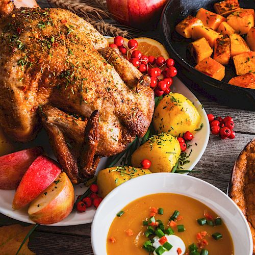 A roasted chicken with potatoes and apples, a bowl of soup with garnish, and a skillet of roasted squash sit on a rustic table.