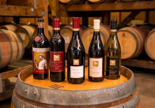 The image shows five bottles of wine placed on a wooden barrel, with wooden barrels in the background, inside what appears to be a winery.