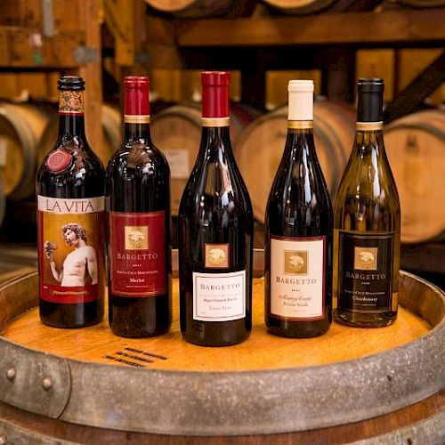 The image shows five bottles of wine placed on a wooden barrel, with wooden barrels in the background, inside what appears to be a winery.