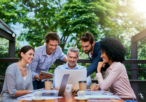 A group of five people, three men and two women, are gathered around a laptop outdoors, smiling and discussing something together.