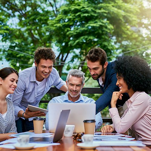 A group of five people, three men and two women, are gathered around a laptop outdoors, smiling and discussing something together.