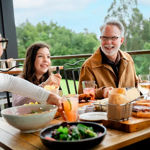 A family sits around a wooden table on a sunny patio, sharing a meal with salads, bowls, bread, and drinks, smiling and chatting.
