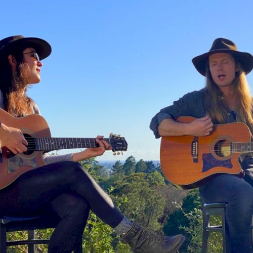 Two people wearing hats are sitting outdoors, playing guitars and singing with a scenic background of trees and a blue sky.