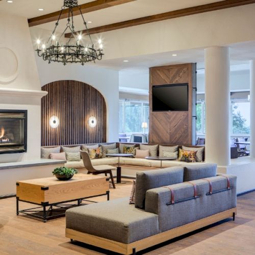 A modern, spacious lounge area with a fireplace, seating, chandelier, and a TV, designed with neutral tones and wooden accents, exudes a cozy ambiance.