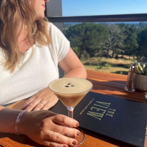 A person is sitting at a table with a cocktail and a menu labeled "The View" against a scenic outdoor backdrop, wearing a white shirt and hat.