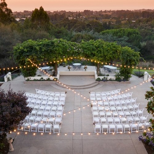 An outdoor wedding venue features a beautifully decorated altar, string lights, rows of white chairs, and lush greenery set against a scenic backdrop.